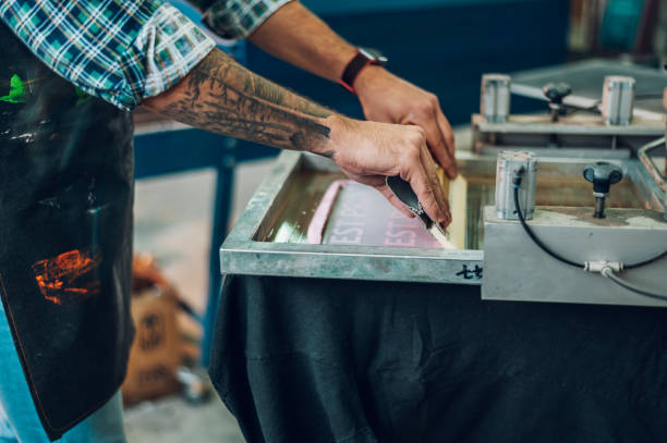 männlicher arbeiter, der tinte auf den rahmen drückt, während er die druckmaschine in einer werkstatt benutzt - silk screen fotos stock-fotos und bilder