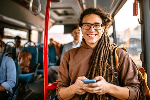 Young smiling student with backpack traveling by bus, using smartphone and headphones while standing in the bus, looking at camera.