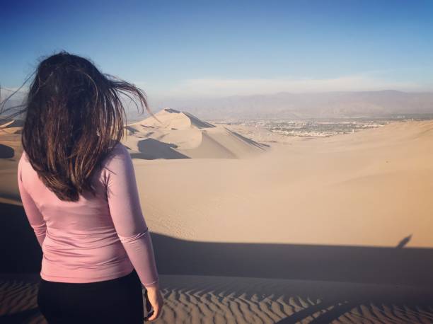 A smiling woman stands barefoot on the rippled sand near the crest of a sunlit dune, holding a tall wooden sandboard upright. She wears a white tank top, rolled blue pants and a red patterned scarf; long shadows and wind-formed sand patterns stretch across the foreground beneath a clear, deep-blue sky.