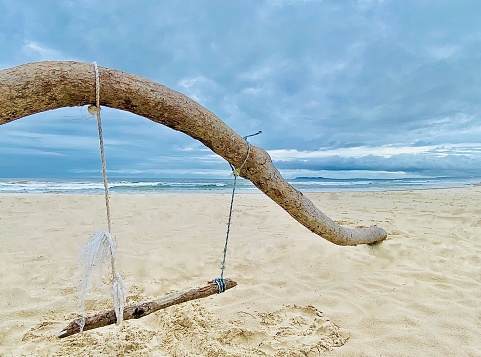 Horizontal seascape of tropical climate driftwood makeshift wood and rope child swing on sand with background breaking waves under a blue stormy overcast cloudscape at Brunswick Heads beach near Byron Bay Australia