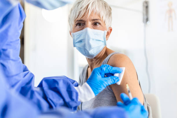 istock Close up of general practitioner hand holding vaccine injection while wearing face protective mask during covid-19 pandemic. Young woman nurse with surgical mask giving injection to senior woman. 1356504930