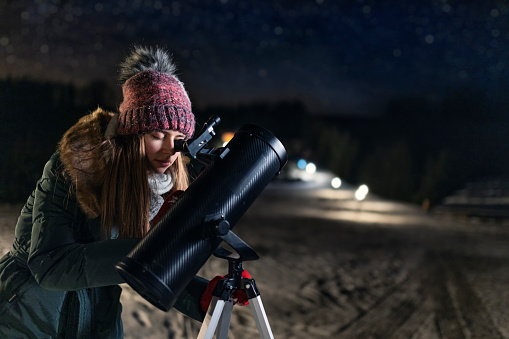 Teenage girl is using the astronomy telescope to observe the the stars at cold winter night.\nShot with Canon R5.