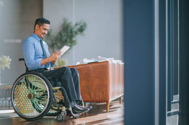 discapacitado hombre indio asiático trabajador de cuello blanco en silla de ruedas leyendo tableta digital sonriendo en la sala de estar - physical impairment wheelchair disabled accessibility fotografías e imágenes de stock