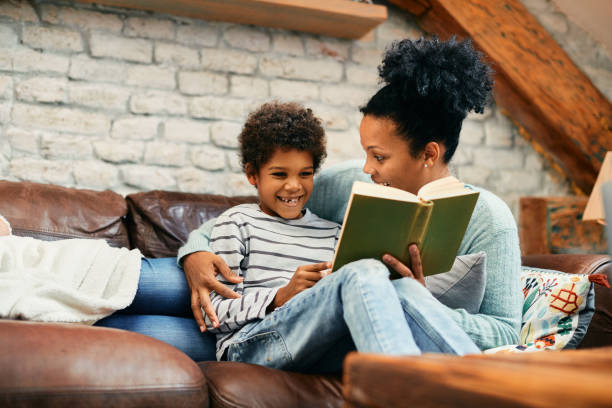 istock Happy African American mother and son reading a book while relaxing on the sofa. 1356510475