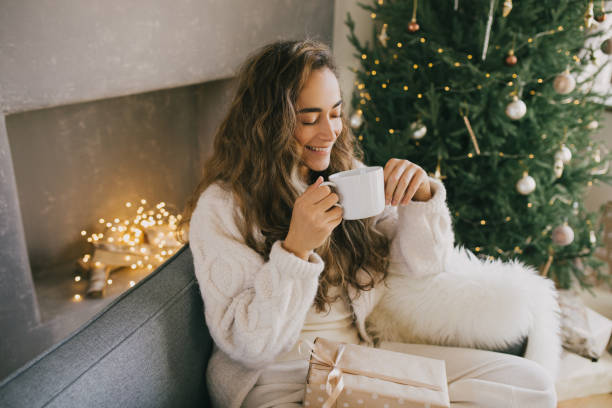 young woman drinking hot coffee or cocoa sitting on couch in cozy room with christmas tree. winter holidays. - hot drink imagens e fotografias de stock