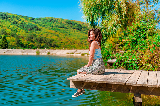 A charming young woman in a skirt is sitting on a wooden bridge against the background of nature with a lake in the forest. Travel, freedom, the concept of an active lifestyle.