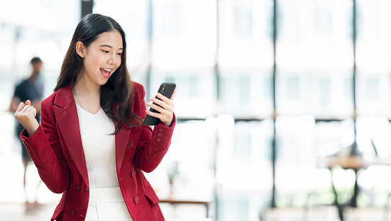 Cheerful asian woman in red suit holding smartphone and celebrate victory triumph while standing in office room.