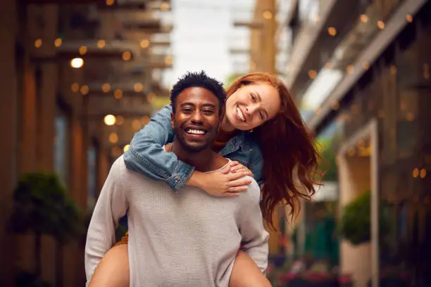 Portrait Of Young Couple Enjoying City Life Heading For Night Out With Man Giving Woman Piggyback