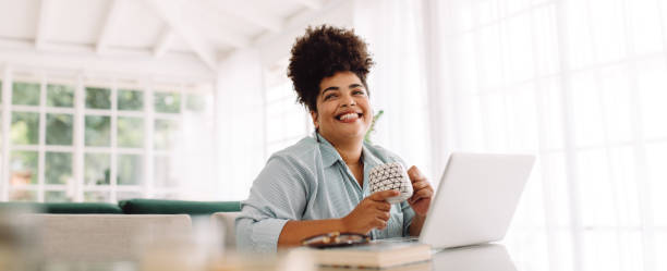 Woman taking break while working from home Wide angle shot of woman sitting on table with coffee mug and laptop. Woman taking break while working from home. happiness photos stock pictures, royalty-free photos & images