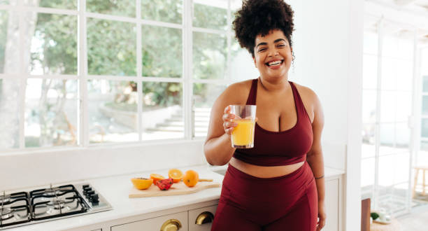 Fitness woman with glass of juice at home Attractive fitness female with glass of fruit juice standing at kitchen. Pretty woman in exercise outfit smiling and holding glass of fruit juice at home. body confidence stock pictures, royalty-free photos & images