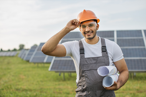 Smiling indian technician in yellow helmet and safety glasses posing at solar station. Worker holding blueprints in hands. Production of green energy.