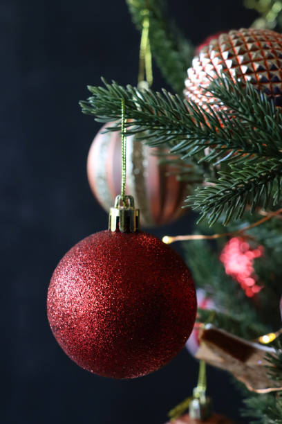 istock Close-up image of individual, red glitter and copper textured baubles hanging on artificial Christmas tree branch, spruce needles, blurred white fairy lights background, copy space 1356532111