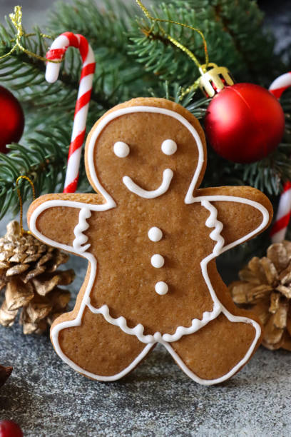 istock Close-up image of gingerbread man biscuit, decorated with white royal icing, leaning against spruce needles of mini Christmas tree, red and gold baubles, grey background, focus on foreground 1356532174