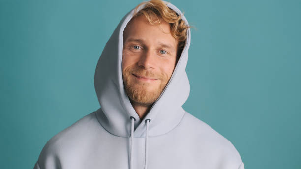 istock Handsome stylish blond bearded guy in hood looking happy smiling on camera over blue background. Attractive casual man 1356543112
