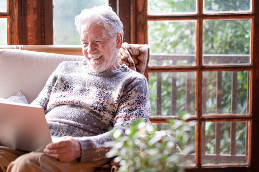 Sonriente hombre mayor de cabello blanco con suéter de invierno sentado en la sala de estar usando una computadora portátil. Abuelo anciano despreocupado que disfruta de la tecnología y las redes sociales. Chalet rústico en madera photo