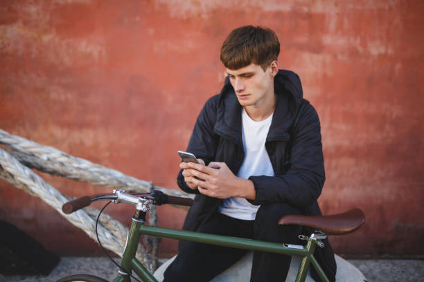 istock Cool boy with brown hair sitting and using his cellphone. Young man in down jacket and white t-shirt sitting with mobile phone in hands and bicycle nearby 1356544838