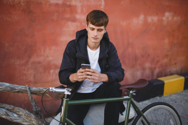 istock Boy with brown hair sitting and using his cellphone. Young thoughtful man in down jacket and white t-shirt sitting with mobile phone in hands and bicycle nearby 1356544858