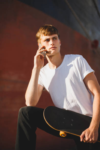 istock Boy with brown hair standing with skateboard in hand and dreamily looking aside while talking on his cellphone. Young thoughtful man in white t-shirt using his mobile phone 1356544968