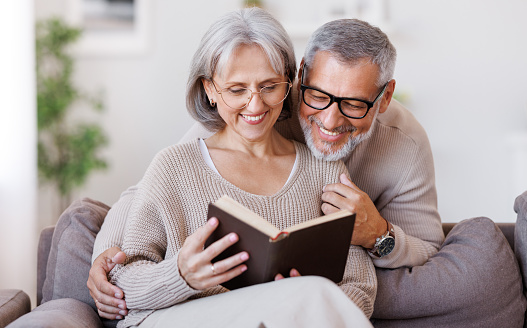 Happy smiling senior family couple in love reading book together, hugging embracing while spending free time at home on retirement, beautiful retired husband and wife enjoying life