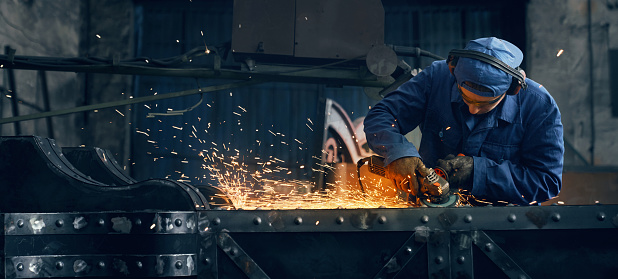 Young man in protective glasses, gloves and headphones sawing metal with special equipment at factory. Sparks flying around. Manufacturing concept.