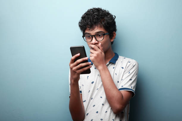 jovem de origem indiana olhando para seu celular com uma expressão de rosto surpreso - só meninos adolescentes - fotografias e filmes do acervo