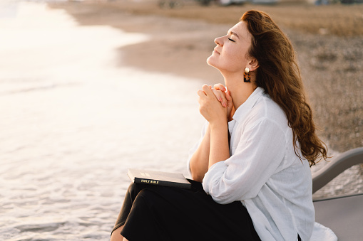Woman closed her eyes, praying on a sea during beautiful sunset. Hands folded in prayer concept for faith, spirituality and religion. Peace, hope, dreams concept