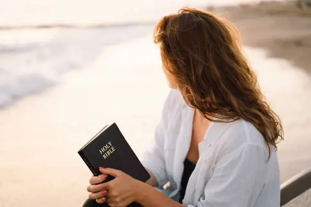 Photo of Christian woman holds bible in her hands. Reading the Holy Bible on the sea during beautiful sunset. Concept for faith, spirituality and religion.