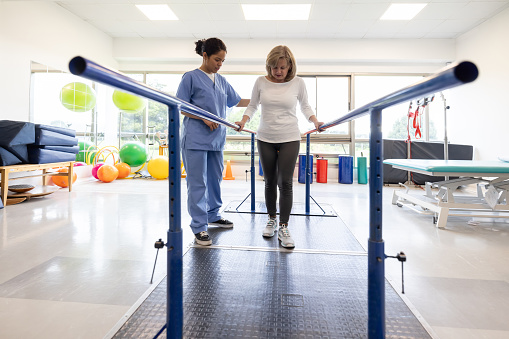 Senior Latin American woman in physiotherapy walking on the parallel bars with he help of her therapist