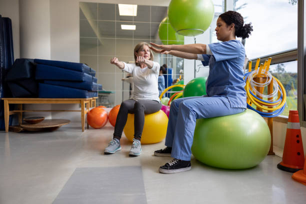 Physical therapist showing a woman an exercise for her recovery Physical therapist showing a Latin American woman a stretching exercise for her recovery at a rehab center while sitting on a fitness ball occupational therapy photos stock pictures, royalty-free photos & images