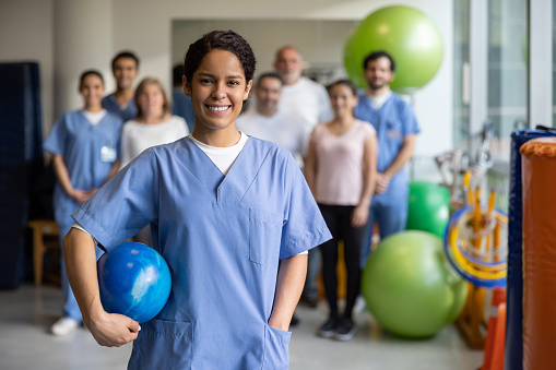 Happy physiotherapist working at a rehab center and smiling in front of a group of coworkers and patients while looking at the camera