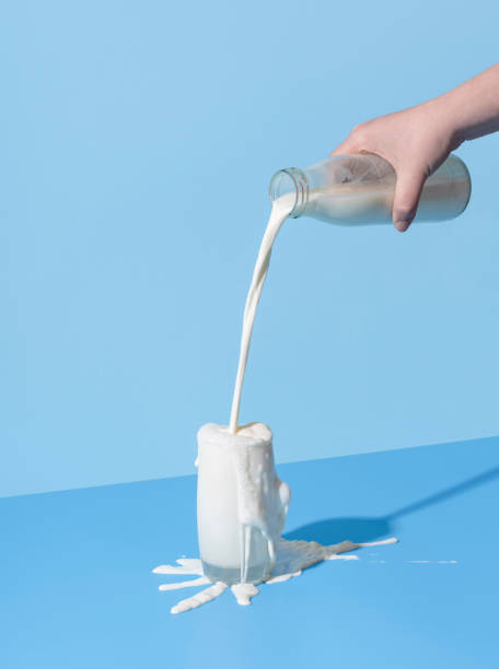 Pouring milk into the glass on a blue background. Spilled milk on the table. Woman hand pouring milk from a bottle into the glass, minimalist on a blue background. Over-spilling milk on the blue-colored table. milk froth stock pictures, royalty-free photos & images