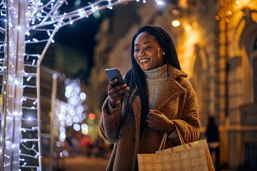 Young smiling african woman standing on the city streets with shopping bags during Christmastime and typing on her cellphone