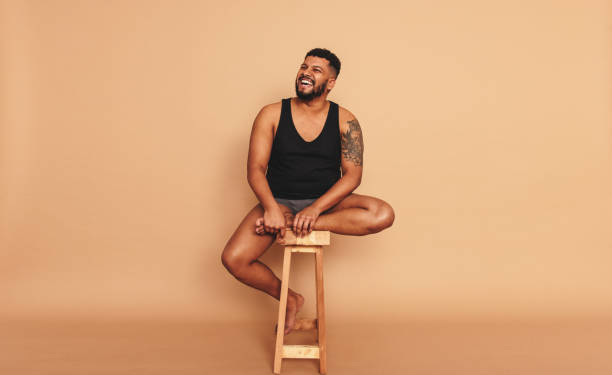 Tattooed man sitting on a wooden chair in a studio Tattooed man smiling cheerfully while sitting on a wooden chair in a studio. Body positive young man looking away while sitting alone. Self-confident young man feeling comfortable in his natural body. body positive stock pictures, royalty-free photos & images