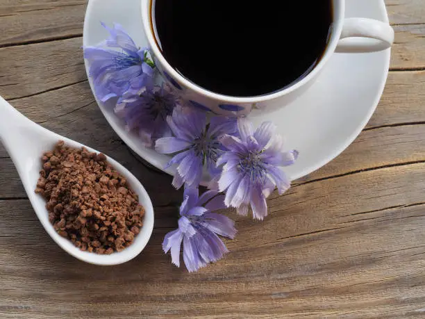 Granulated sublimated chicory in a white spoon, blue flower and chicory drink in a white сup on a wooden background, top view. Healthy natural coffee from the Cichorium plant
