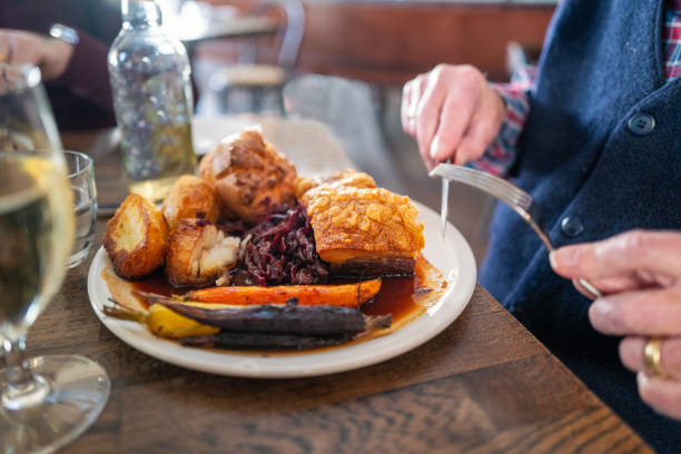The golden thick crackling on top of roast pork belly on a white plate with roast potatoes and Yorkshire pudding with vegetables in a restaurant. A mans hands can be seen holding a knife and fork. The golden thick crackling on top of roast pork belly on a white plate with roast potatoes and Yorkshire pudding with vegetables in a restaurant. A mans hands can be seen holding a knife and fork. roast dinner stock pictures, royalty-free photos & images