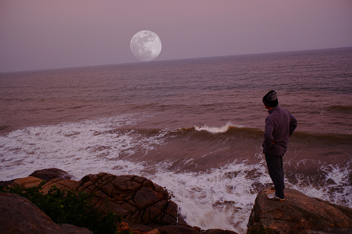 Man in silhouette looking at the distance at the full moon overseas with a calm orange sky.