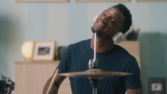 Young African American man listening to rhythmic music in wireless headphones and playing drums during rehearsal in home studio in daytime