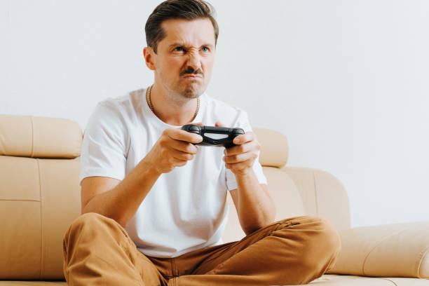 Angry guy gamer playing video game using wireless joystick while sitting on sofa at home. Aggressive, loser young man player, irritated with defeat in e-sports
