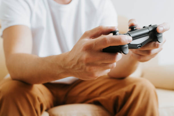 istock Close-up of gamer's male hands holding wireless game controller with joysticks. Side view of man fun, playing video game on console while sitting on couch at home. Cropped, selective focus on front 1356590308