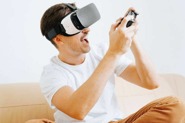 istock Cheerful young man with wow expression in 3d glasses or vr helmet holding wireless gamepad controller and playing virtual video game while sitting on sofa. Side view of guy player enjoying hobby 1356590320
