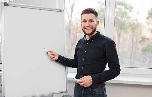 Smiling male employee pointing at blank whiteboard and explaining business strategy in workplace while looking at camera