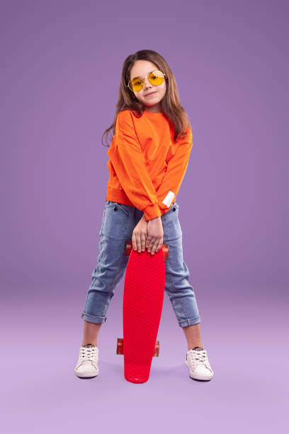 Stylish little girl with skateboard Positive active hipster little girl in trendy outfit and eyewear holding red skateboard and looking at camera on purple background young cool girl stock pictures, royalty-free photos & images