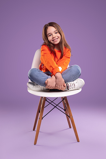 joyful little girl in orange sweater looking at camera with curiosity while sitting on chair with arms and legs crossed against violet background in studio