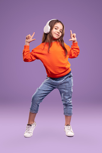 Full body positive child in casual outfit pointing up and looking looking at camera while listening to music against violet background