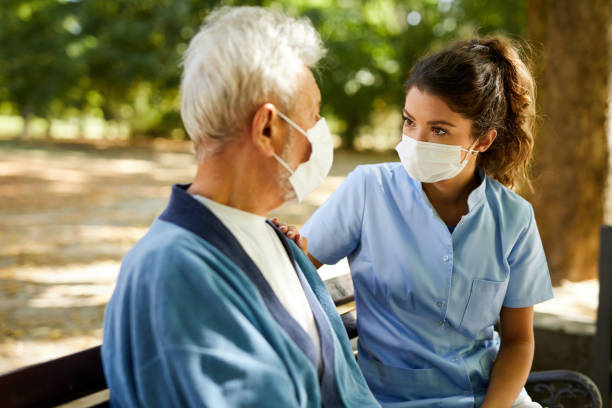 Healthcare worker and senior man with protective face masks on sitting on bench and communicating stock photo