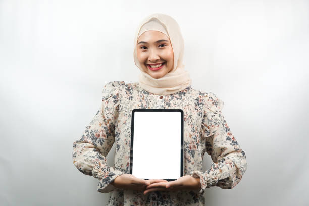 Beautiful young asian muslim woman smiling, excited and cheerful holding tablet with white/blank screen, promoting app, promoting product, presenting something, isolated on white background stock photo