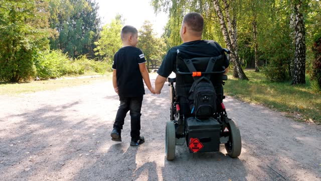 father's day. person with disability. dad and his little cute son are walking in park, on summer day. dad is a wheelchair user. disabled people. people with special needs