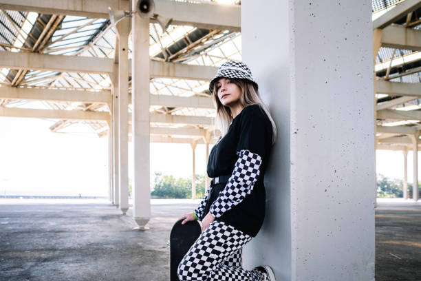 Stylish millennial woman with skateboard leaning on pillar Side view of serious young woman in casual stylish wear standing with skateboard near concrete column in abandoned building alternative rock stock pictures, royalty-free photos & images