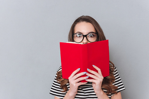 Picture of surprised young lady standing over grey wall holding book. Looking at camera.