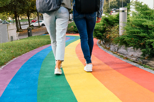 Rear cropped view of the legs of two women walking along a road painted with the colors of the rainbow.